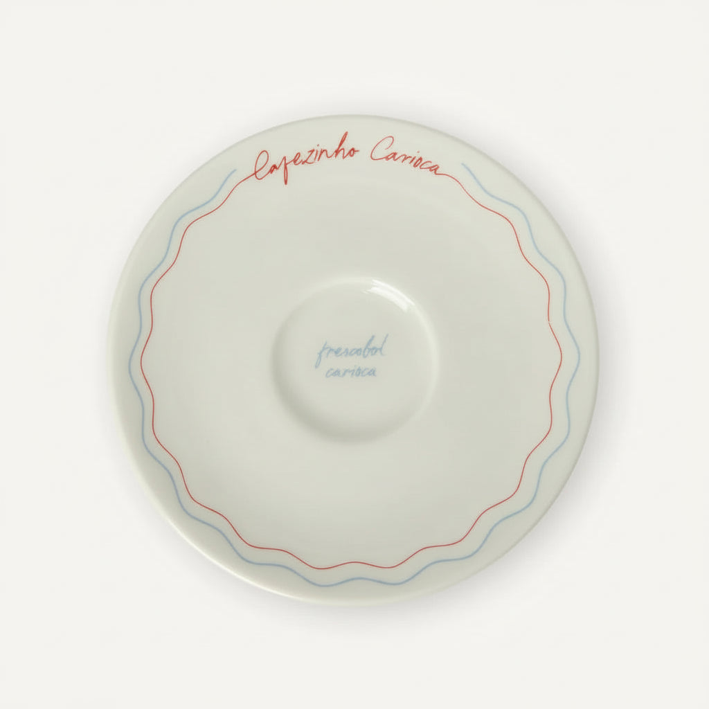 A white ceramic coffee saucer with a scalloped rim, featuring a double line of wavy red and blue detailing and handwritten text in red and blue.