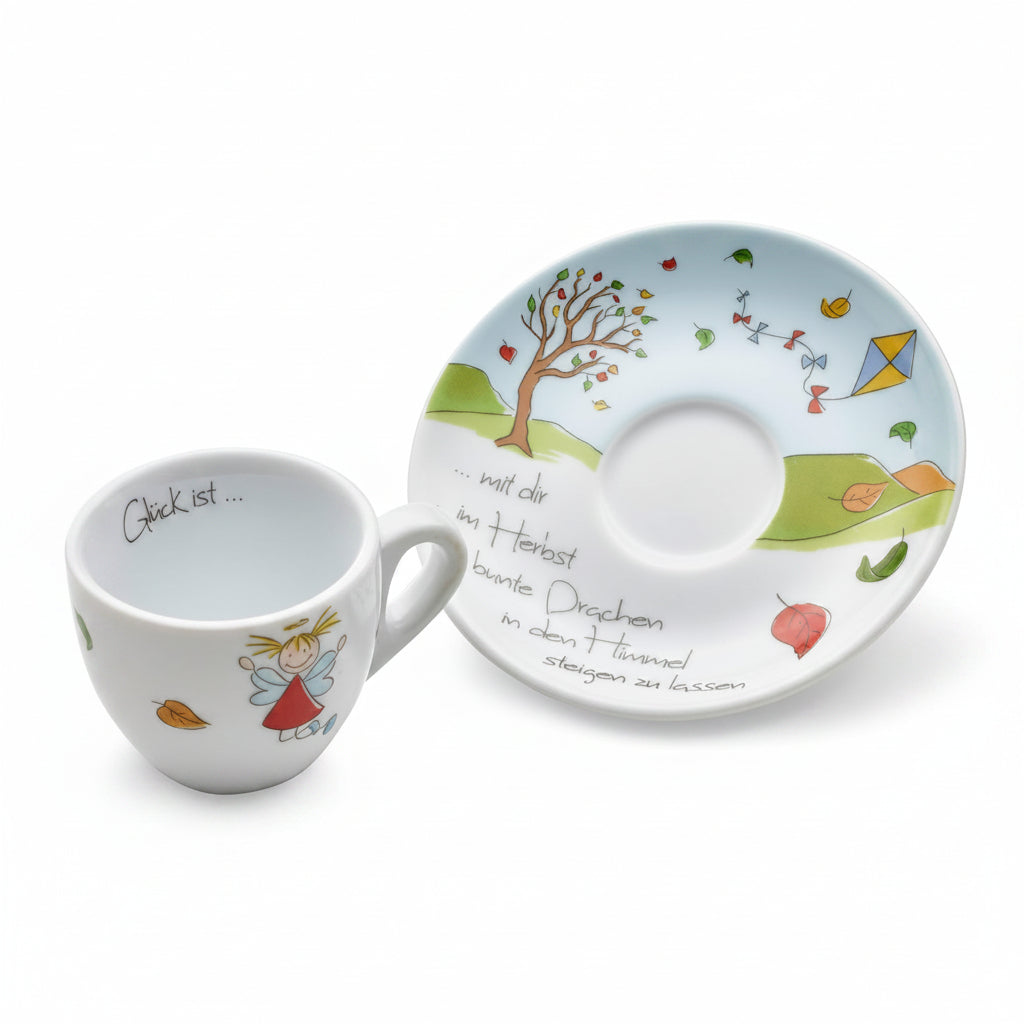 A white ceramic cup and matching saucer set with whimsical hand-painted illustrations of a tree, hills, leaves, a kite, and a fairy, along with German text.