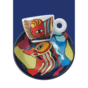 A white ceramic espresso cup and matching saucer set, both decorated with a colorful abstract face illustration in red, yellow, blue, and cream, set against a dark blue background.