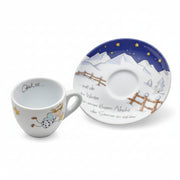A white ceramic cup and matching saucer decorated with a winter night scene, featuring a blue sky with yellow stars, snowy mountains, wooden fences, and a small angel figure. The cup has German text inside, and the saucer has German text on the side.