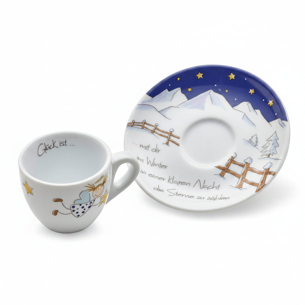 A white ceramic cup and matching saucer decorated with a winter night scene, featuring a blue sky with yellow stars, snowy mountains, wooden fences, and a small angel figure. The cup has German text inside, and the saucer has German text on the side.