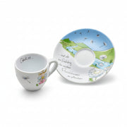 A white ceramic cup and matching saucer set with a whimsical spring landscape illustration, featuring a blue sky, green hills, a winding river, dandelions, and German text on the saucer.